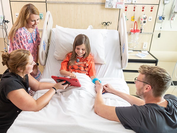 pediatric patient lying in bed being entertained by a laptop held by a child life specialist while mom watches and another provider puts something on her arm