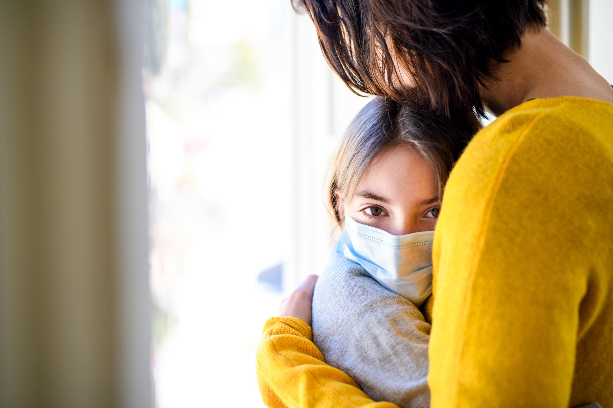 Small schoolgirl with face mask and unrecognizable mother indoors at home, hugging.