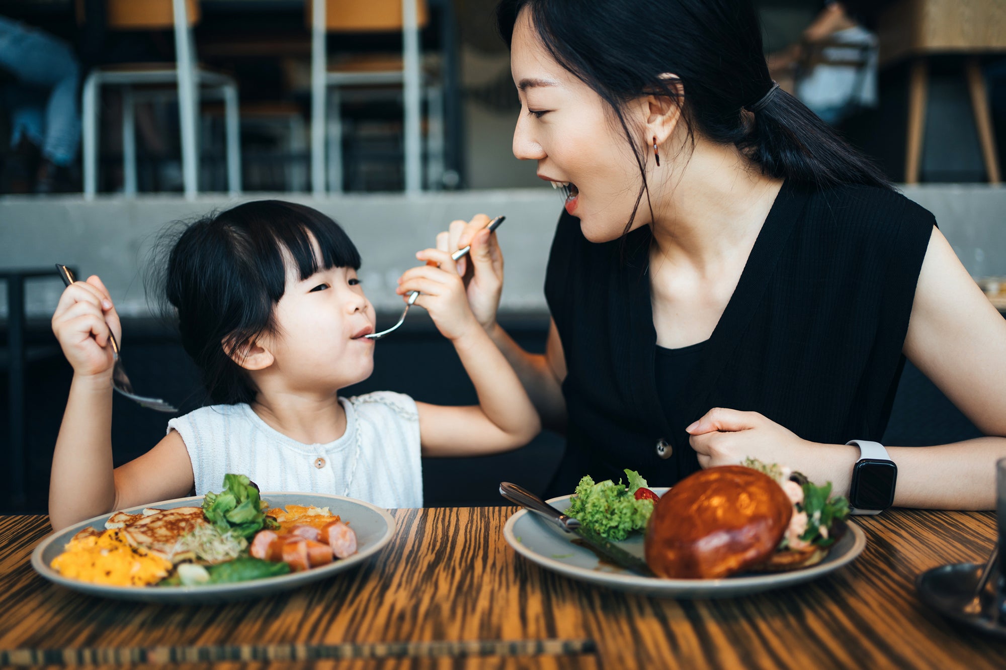 Young Asian family having lunch in cafe, mother sharing food to her little daughter and enjoying a happy meal together. Family and eating out lifestyle